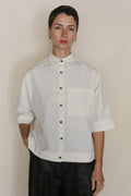 A person with short dark hair wears the Work Shirt Crop | Undyed, a handmade cropped shirt crafted from organic typewriter cotton with short sleeves and a large chest pocket, standing against a neutral background.
