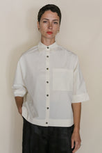 Load image into Gallery viewer, A person with short dark hair wears the Work Shirt Crop | Undyed, a handmade cropped shirt crafted from organic typewriter cotton with short sleeves and a large chest pocket, standing against a neutral background.