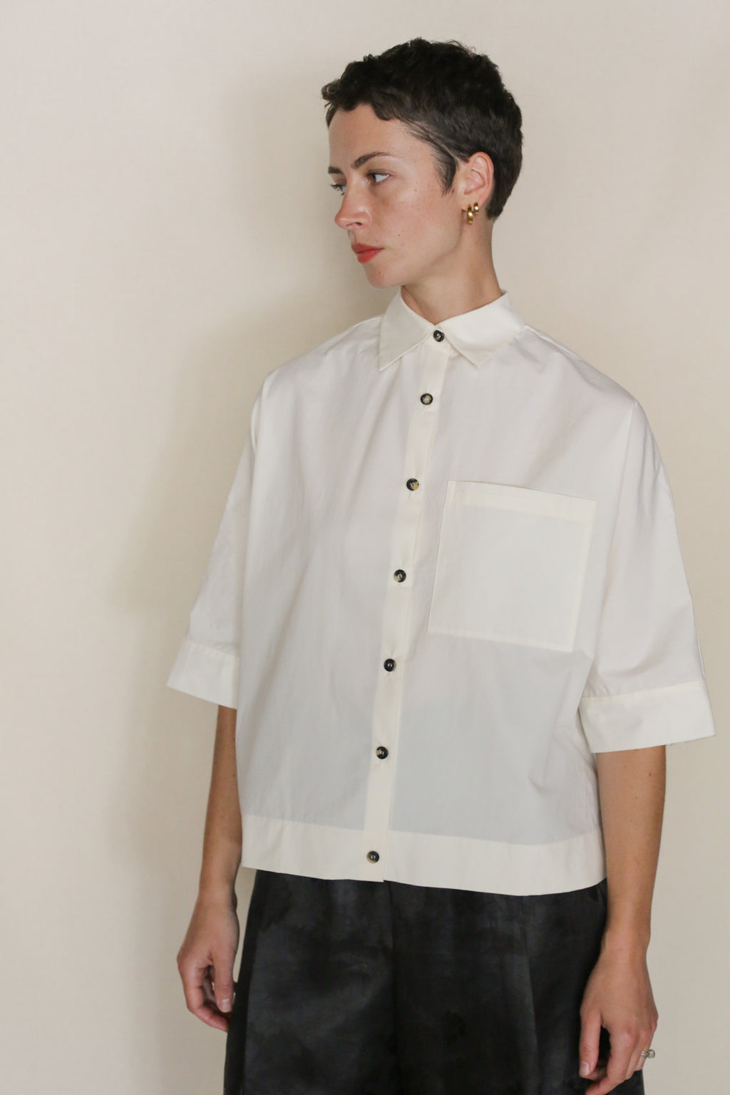 A person with short dark hair, wearing the Work Shirt Crop | Undyed—a cropped shirt made from organic typewriter cotton with a large front pocket and black buttons—stands against a plain light background, looking to the side neutrally.