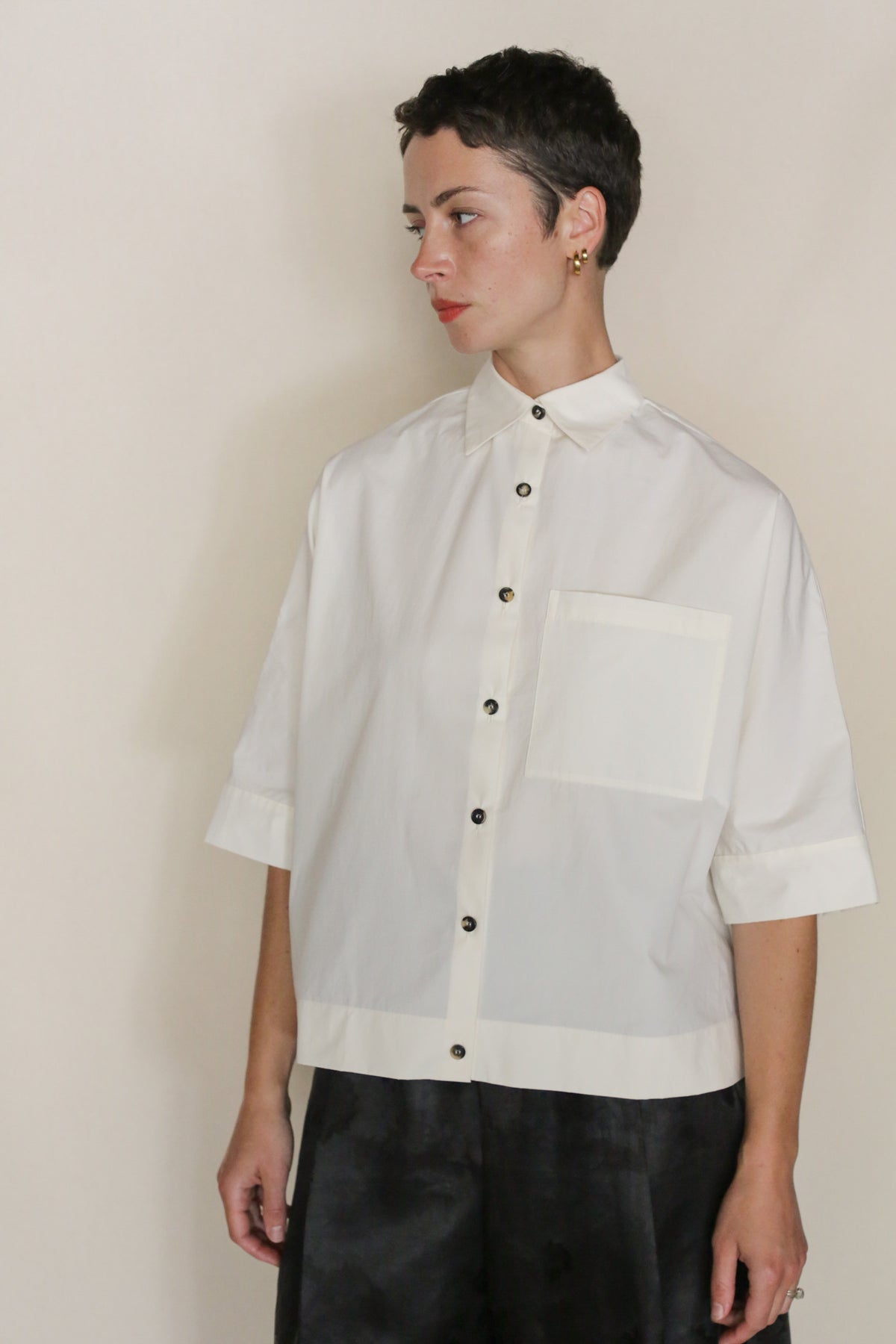 A person with short dark hair, wearing the Work Shirt Crop | Undyed—a cropped shirt made from organic typewriter cotton with a large front pocket and black buttons—stands against a plain light background, looking to the side neutrally.
