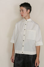 Load image into Gallery viewer, A person with short dark hair, wearing the Work Shirt Crop | Undyed—a cropped shirt made from organic typewriter cotton with a large front pocket and black buttons—stands against a plain light background, looking to the side neutrally.