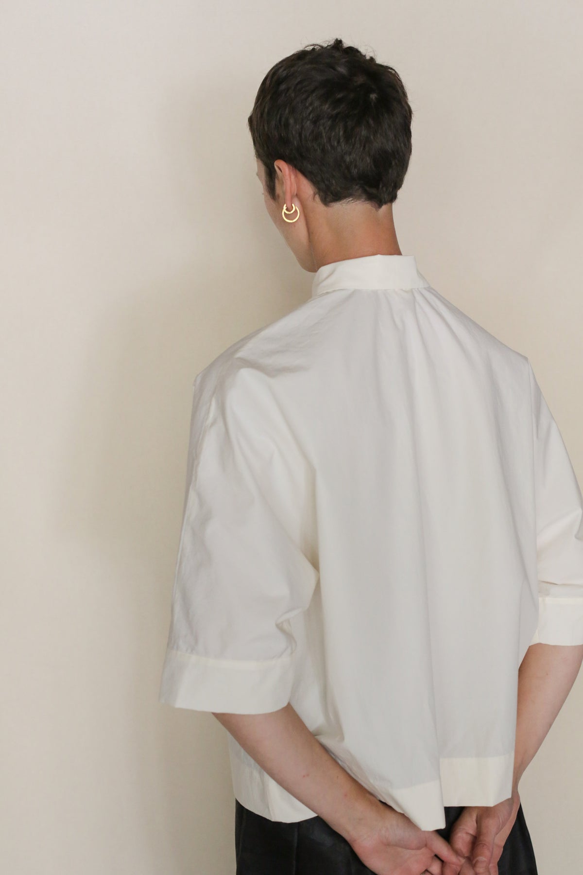 A person with short dark hair wears the Work Shirt Crop | Undyed—an organic typewriter cotton, loose-fit cropped shirt with short sleeves and a stand collar. Facing away, hands behind their back, a gold hoop earring shines on their left ear.