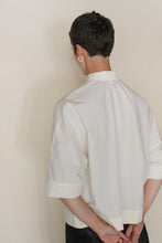 Load image into Gallery viewer, A person with short dark hair wears the Work Shirt Crop | Undyed—an organic typewriter cotton, loose-fit cropped shirt with short sleeves and a stand collar. Facing away, hands behind their back, a gold hoop earring shines on their left ear.