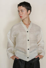 Load image into Gallery viewer, A person with short dark hair and gold hoop earrings wears the Sumi Top | Silk Mesh—a sheer white top with black buttons—standing hands in pockets, looking to the side against a plain background.