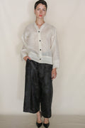 A person stands against a plain background wearing the Silk Mesh Sumi Top with black buttons, paired with wide-leg black pants and pointed shoes—a modern look featuring this lightweight, textured silk mesh cover-up.