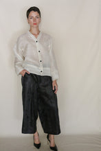 Load image into Gallery viewer, A person with short dark hair poses against a plain light background, wearing the Sumi Top | Silk Mesh—a loose, sheer white button-up shirt—paired with wide-leg black pants and black heels, one hand in a pocket.