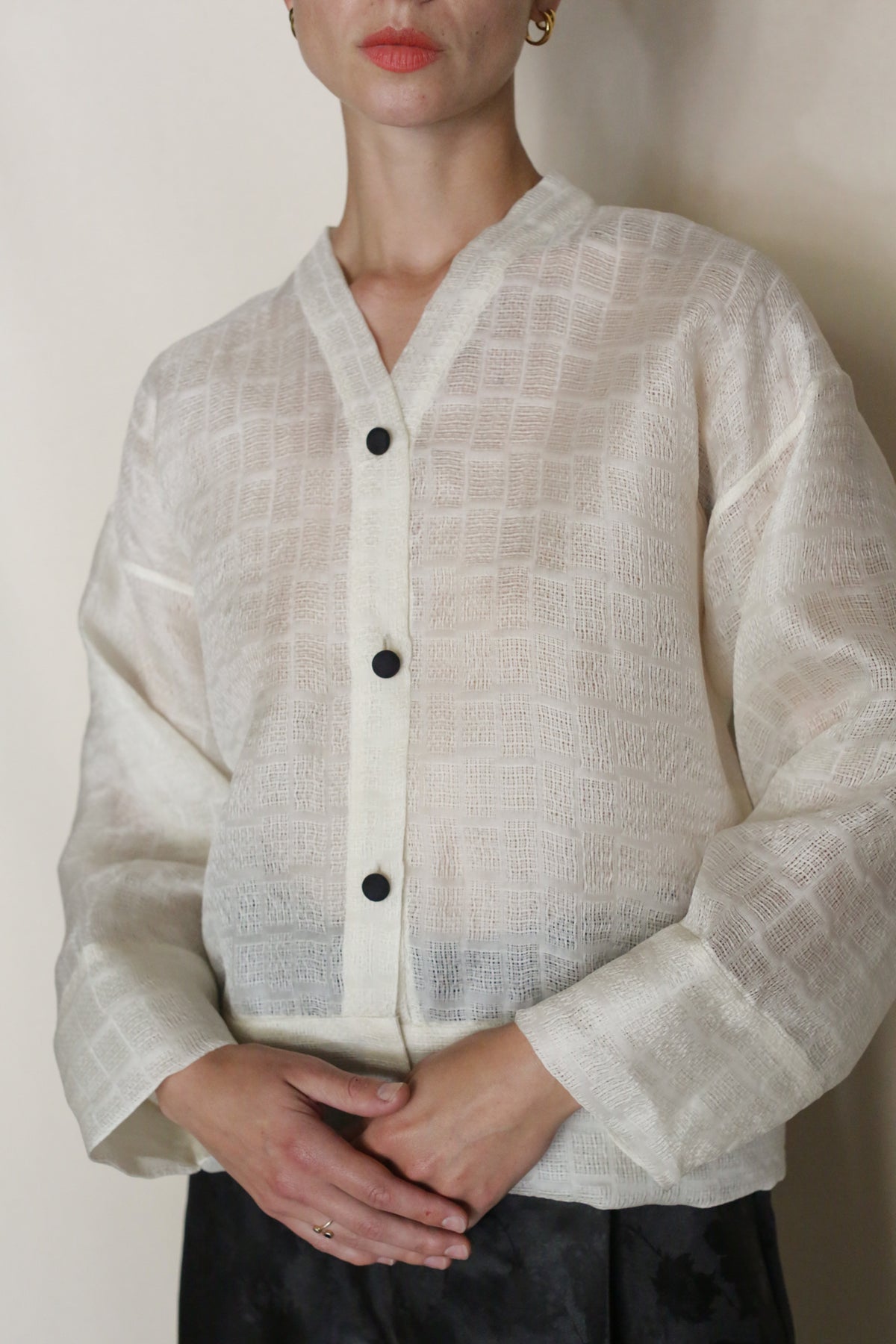 A woman stands against a neutral background, wearing the Silk Mesh Sumi Top—a sheer, textured silk mesh blouse with black buttons—paired with gold hoop earrings, her hands gently clasped at her waist.