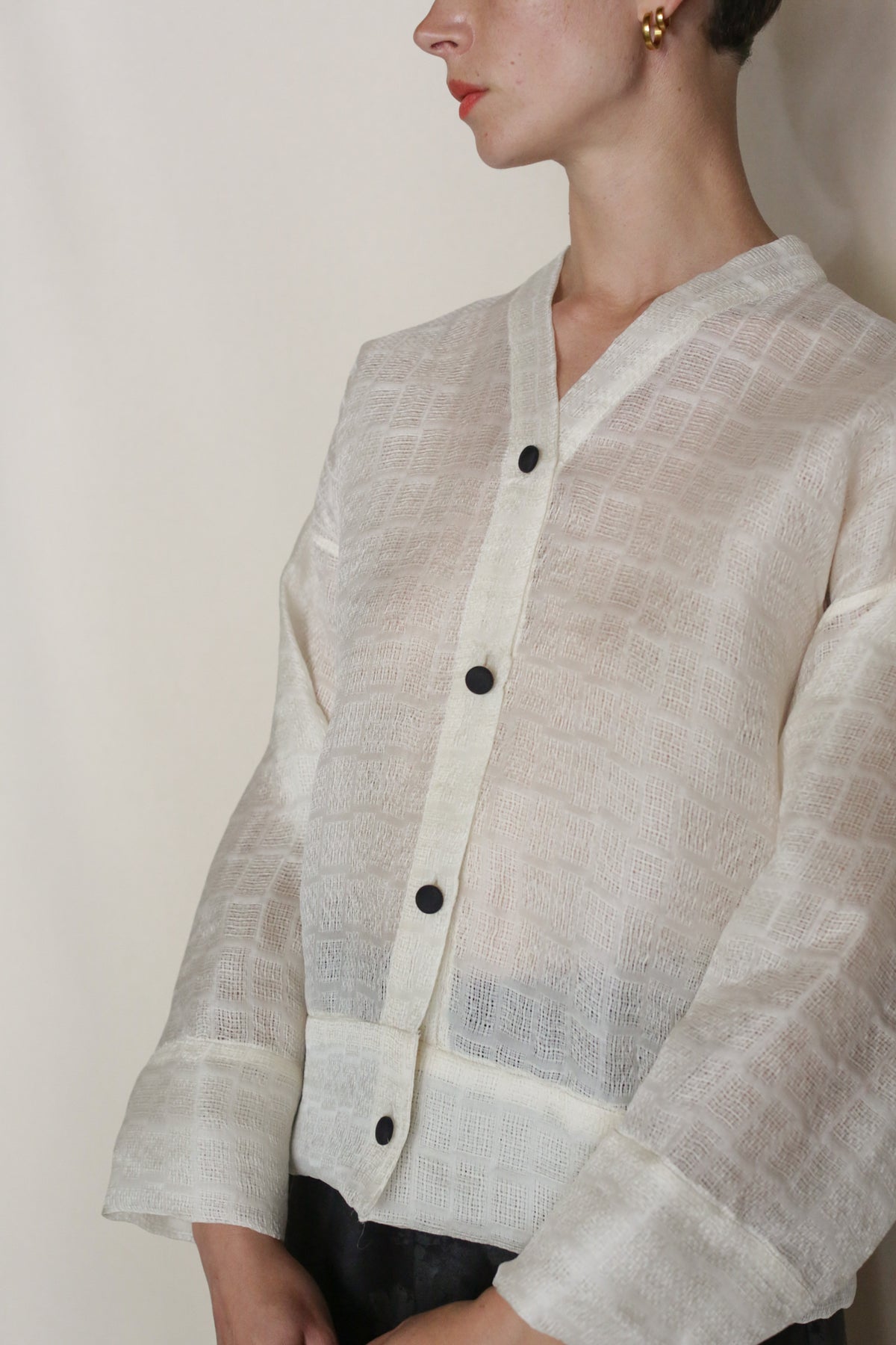 A person wears the Silk Mesh Sumi Top—a sheer, textured silk mesh piece with black buttons and wide sleeves—paired with gold hoop earrings and red lipstick, shown from the shoulders up against a plain background; handmade in San Francisco.