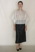 A woman with short hair poses against a plain background, wearing a sheer white blouse with wide sleeves, the Endless Bloom Futo Skirt in black, and black pointed-toe heels.