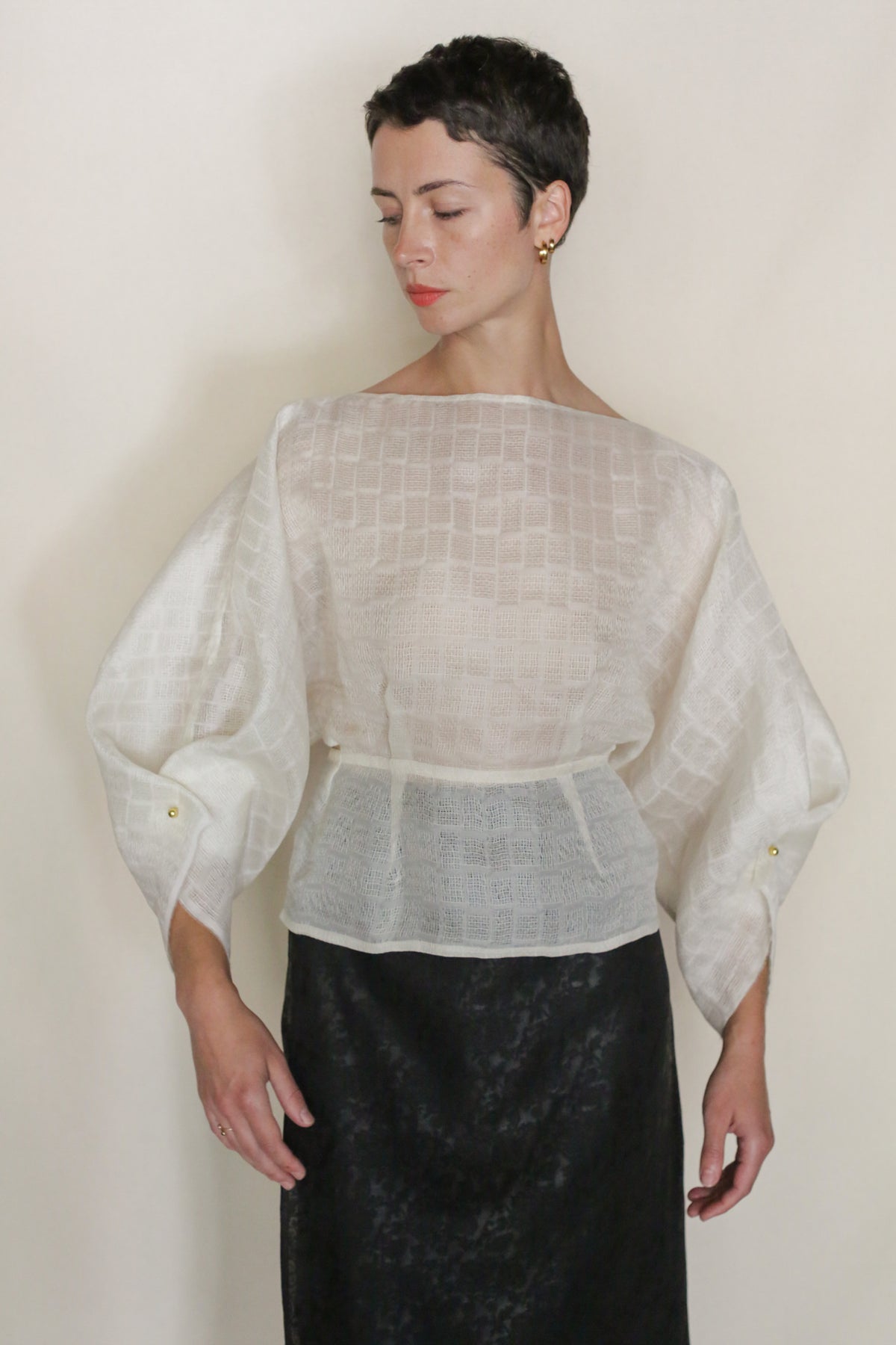 A person with short dark hair wears the Silk Mesh Origami blouse—an origami-inspired sheer white long-sleeved top with gold cuff buttons—paired with a high-waisted black skirt, standing against a plain light background.