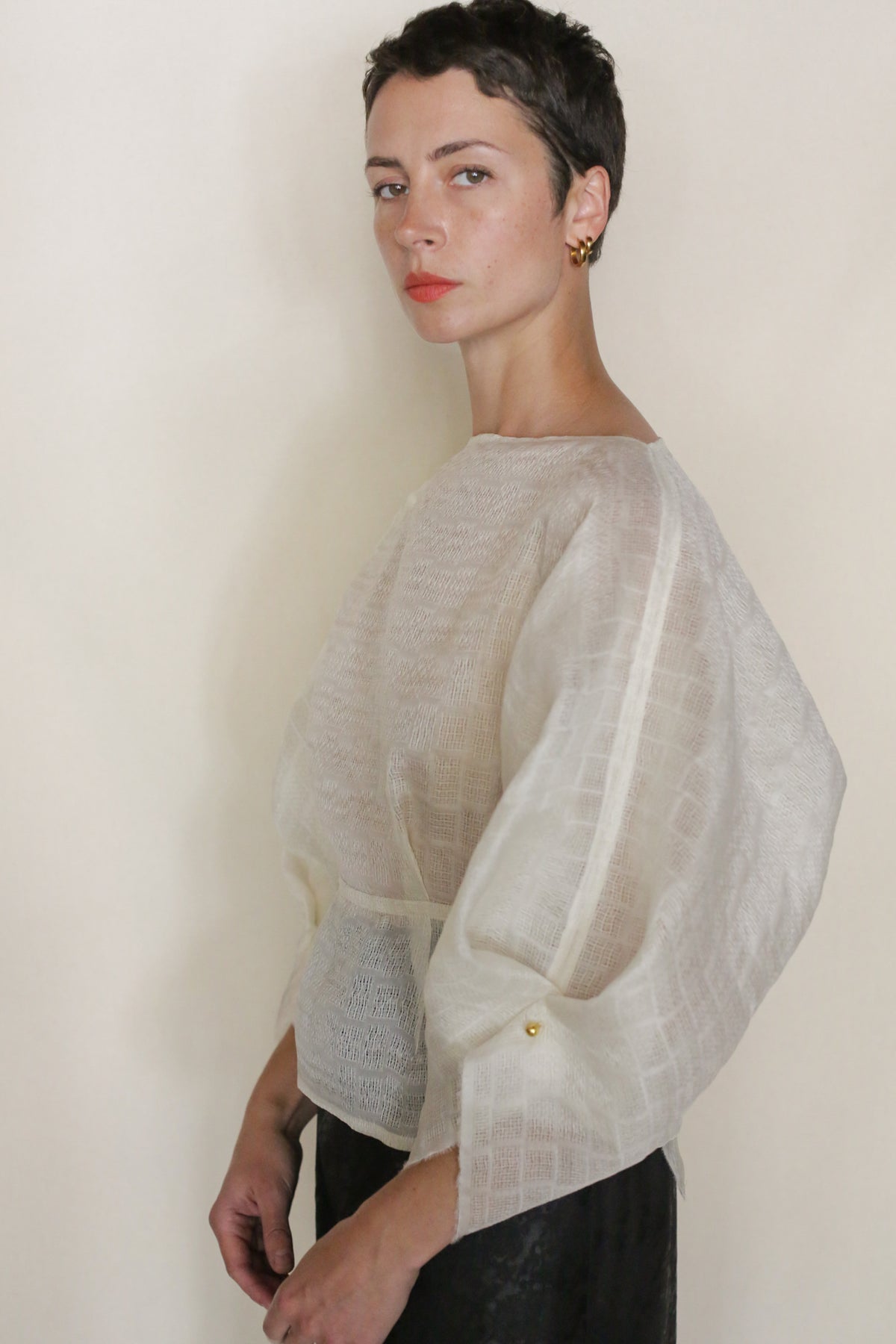 A person with short dark hair and gold earrings stands sideways, thoughtfully looking at the camera in the Silk Mesh Origami top with wide sleeves and a black skirt against a plain background.
