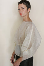 Load image into Gallery viewer, A person with short dark hair and gold earrings wears the Origami | Silk Mesh top with wide sleeves, standing against a plain light background and gazing seriously at the camera.