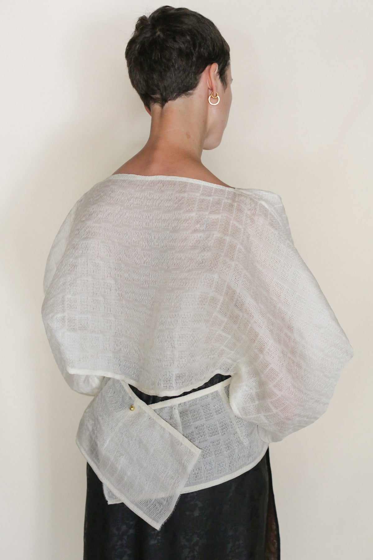 A person with short dark hair, seen from behind, wears the Silk Mesh Origami—a textured white zero-waste shawl with a buttoned square pouch at the lower back—over a black outfit, against a plain light background.