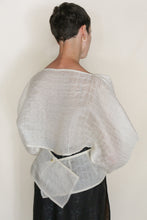 Load image into Gallery viewer, A person with short dark hair, seen from behind, wears the Origami | Silk Mesh—a sheer white grid-patterned shawl inspired by zero-waste design, featuring a square fabric detail pinned at the lower back over a black outfit.