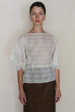 Load image into Gallery viewer, A person with short dark hair wears the Origami | Silk Double Cloth Woven Veil blouse, featuring sheer white fabric and half-length puff sleeves, paired with a brown skirt against a plain light background.