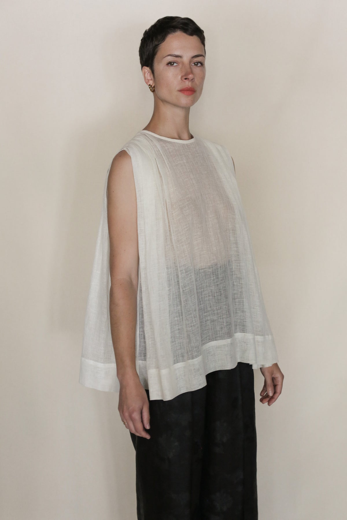 A person with short dark hair wears the handmade Leno Pleat Top—a sleeveless, loose, semi-sheer linen piece—over black pants against a plain light background. This San Francisco design adds effortless style to any look.