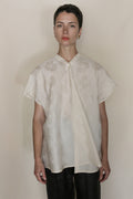 A person with short dark hair wears the Hidden Peony Qipao Vest in light beige silk-cotton jacquard with a subtle floral pattern and black pants, standing against a plain light background.