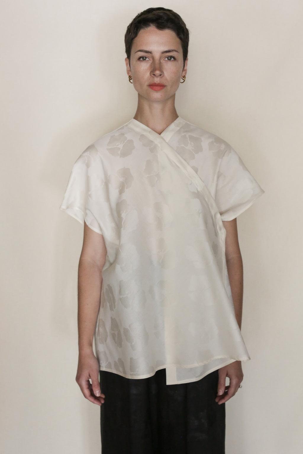 A person with short dark hair faces forward, wearing the handmade Hidden Peony Qipao Vest in cream silk-cotton jacquard with a subtle floral pattern and dark pants, against a plain light background.