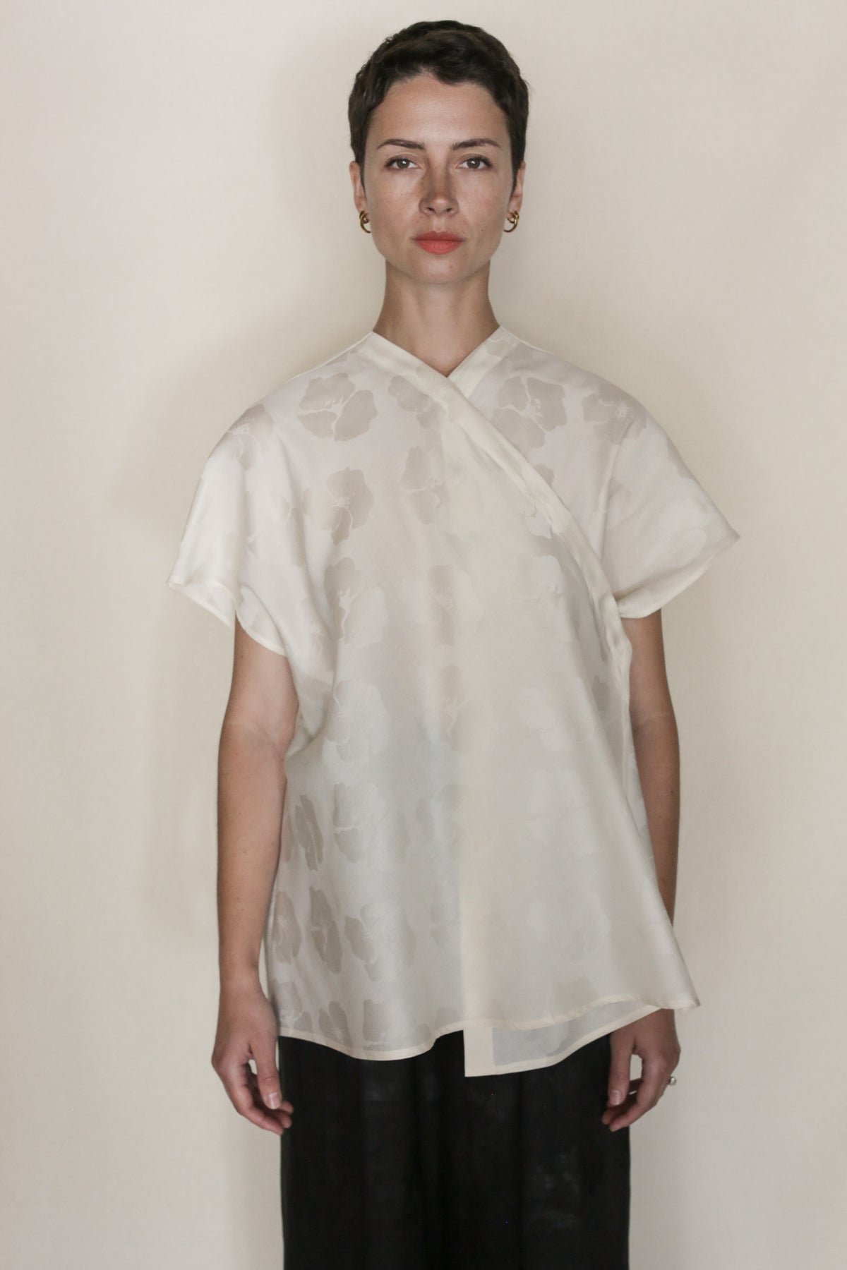 A person with short dark hair faces forward, wearing the handmade Hidden Peony Qipao Vest in cream silk-cotton jacquard with a subtle floral pattern and dark pants, against a plain light background.