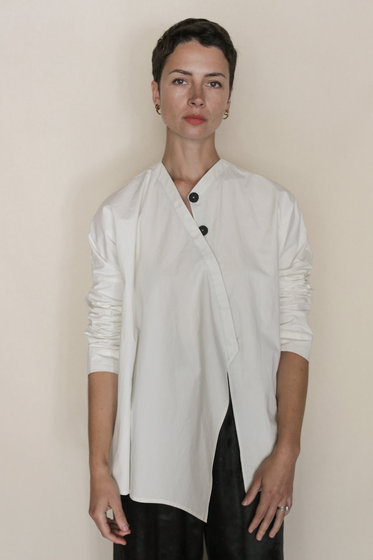 A person with short dark hair stands against a light background, wearing the handmade Omizarashi Qipao Blouse with black buttons and dark pants. Their expression is neutral, arms relaxed by their sides.