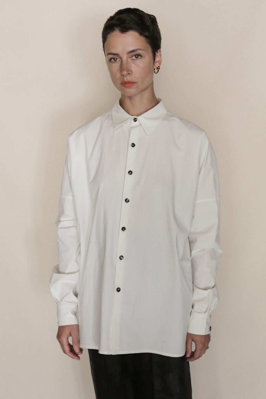 A person stands against a neutral background, wearing the Omizarashi Unisex Shirt | White—an oversized shirt crafted from luxurious Ohmi Zarashi cotton with dark buttons—paired with black pants. They have short dark hair, a neutral expression, and their distinctive shirt subtly reflects refined handmade San Francisco craftsmanship.