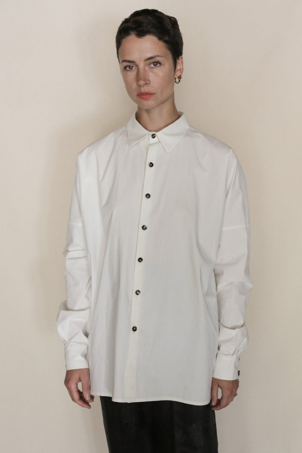 A person stands against a neutral background, wearing the Omizarashi Unisex Shirt | White—an oversized shirt crafted from luxurious Ohmi Zarashi cotton with dark buttons—paired with black pants. They have short dark hair, a neutral expression, and their distinctive shirt subtly reflects refined handmade San Francisco craftsmanship.