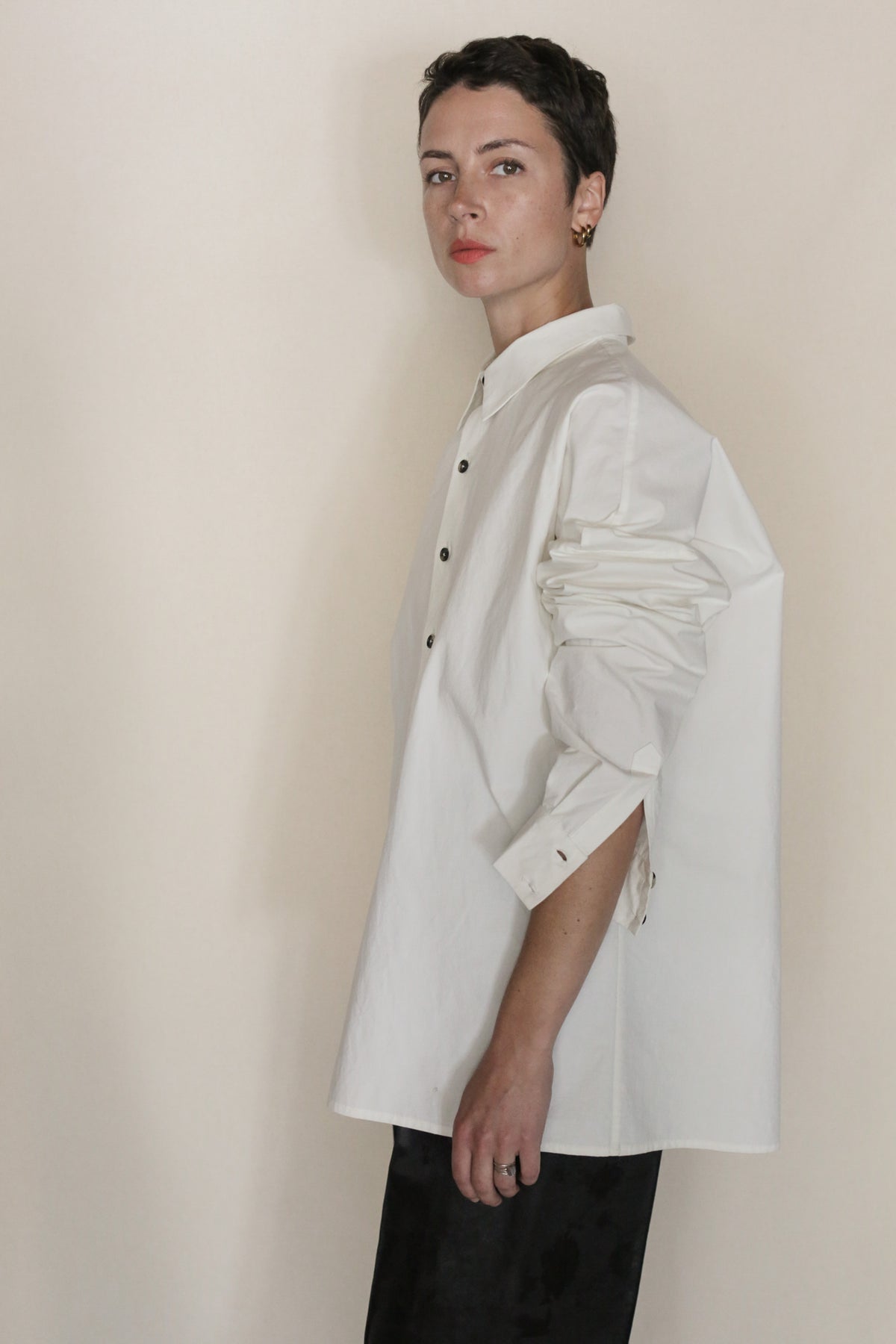 A person with short hair models the Omizarashi Unisex Shirt in white—crafted from Ohmi Zarashi cotton—and dark pants, standing sideways and looking at the camera against a plain, light-colored background. Handmade in San Francisco, this shirt radiates effortless style and comfort.