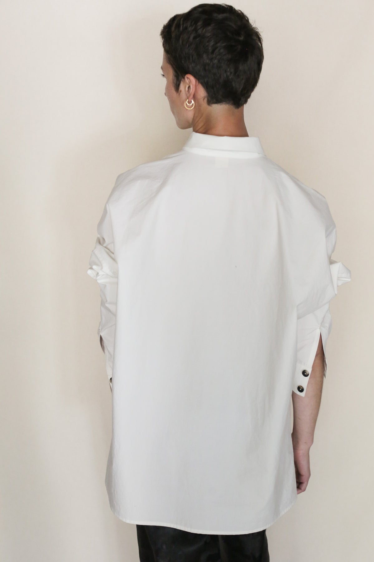 A person stands with their back to the camera, wearing the Omizarashi Unisex Shirt in white—a handmade button-up crafted from Ohmi Zarashi cotton with rolled-up sleeves—paired with black pants, set against a plain light background.