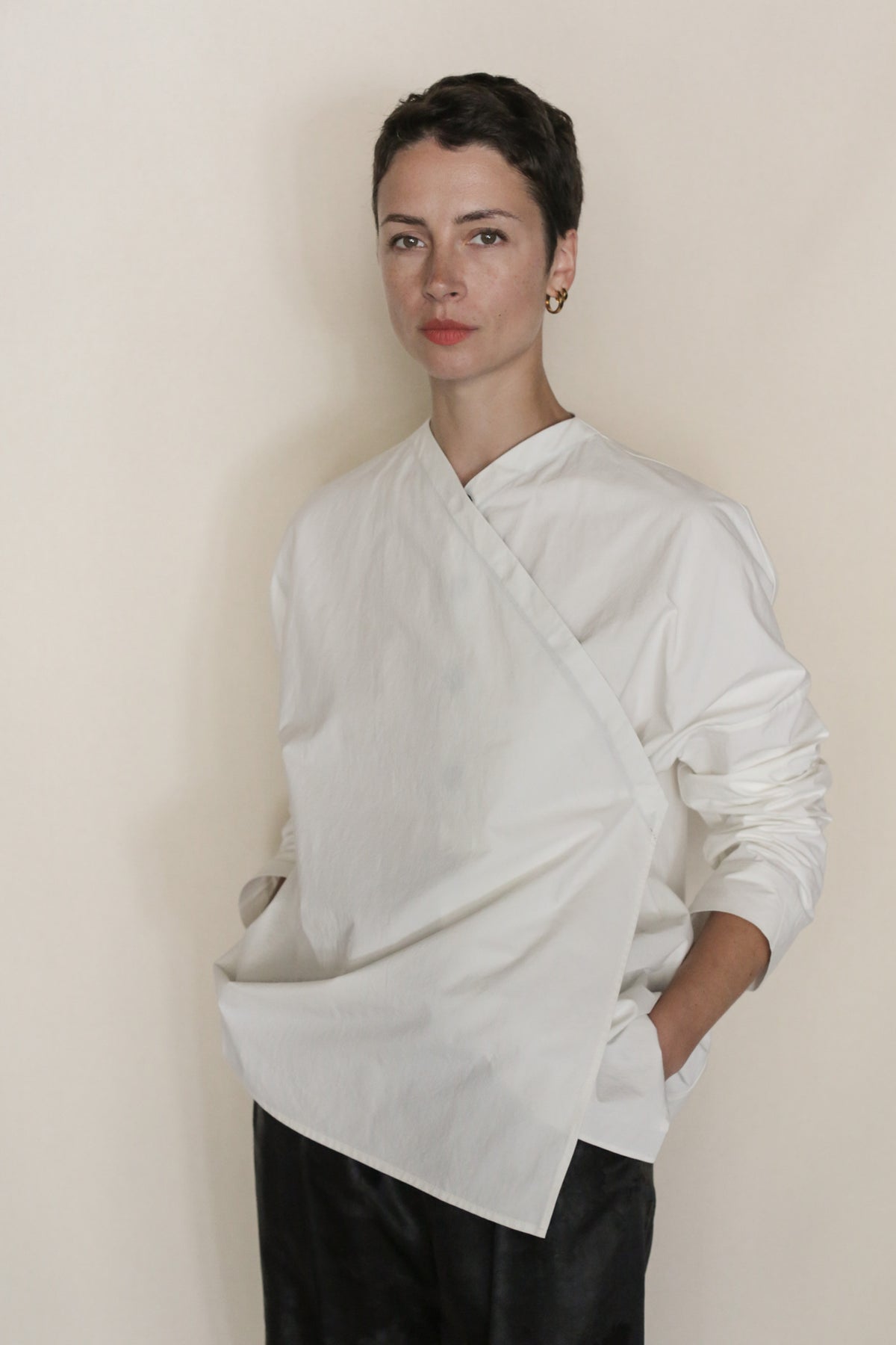 A person with short dark hair and a neutral expression wears an Omizarashi Qipao Blouse—a handmade, long-sleeved white top of Ohmi Zarashi cotton with wide panels—and dark pants, standing against a plain light background.