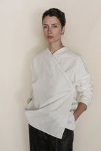 Load image into Gallery viewer, A person with short dark hair, wearing a loose-fitting, asymmetrical Qipao Blouse | Cotton Omizarashi Undyed and black pants, stands against a plain light background with hands in pockets, looking calmly at the camera.