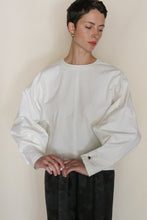 Load image into Gallery viewer, A person with short dark hair wears the Cocoon | Cotton Omizarashi Undyed—a loose, long-sleeved white cotton top—with dark pants, standing against a plain background and looking down with a neutral expression.