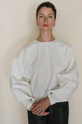 A person with short dark hair and hoop earrings wears the Omizarash Cocoon, a loose-fitting, long-sleeved white top crafted from heritage Japanese cotton, standing against a plain background with hands clasped, looking at the camera.