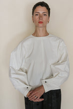 Load image into Gallery viewer, A person with short dark hair wears the Cocoon | Cotton Omizarashi Undyed, a loose-fitting white long-sleeve top made from renowned Japanese Ohmi Zarashi cotton, standing against a plain light background with hands clasped and a neutral expression.