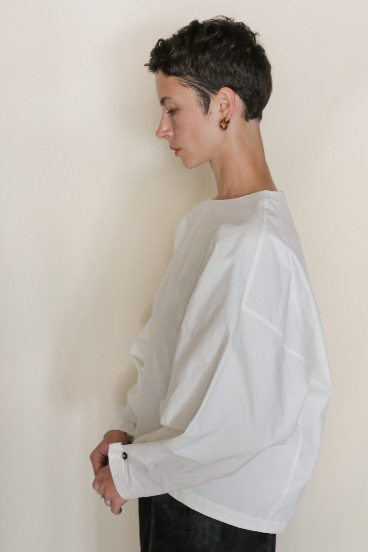 A woman with short dark hair, gold hoop earrings, and a loose white Omizarash Cocoon top—handmade in San Francisco—stands in profile against a plain light background with her hands clasped in front of her.