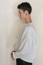 Load image into Gallery viewer, A woman with short dark hair stands in profile against a plain background, wearing the Cocoon | Cotton Omizarashi Undyed blouse with wide sleeves and gold hoop earrings, looking down with her hands clasped in front of her.