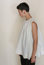 Load image into Gallery viewer, A person with short dark hair stands sideways, looking down, wearing the Shoulder Pleat Top | Omizarashi Undyed— a loose, sleeveless white top— and black pants against a plain light background.