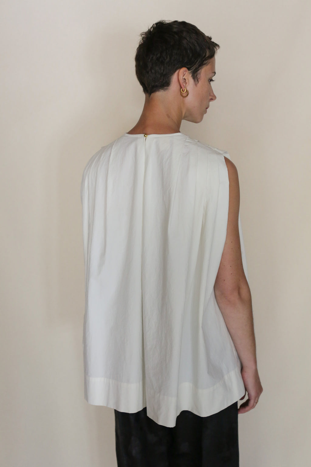 A person with short hair, wearing the Omizarashi Pleat Top and black pants, stands facing away from the camera against a neutral background.
