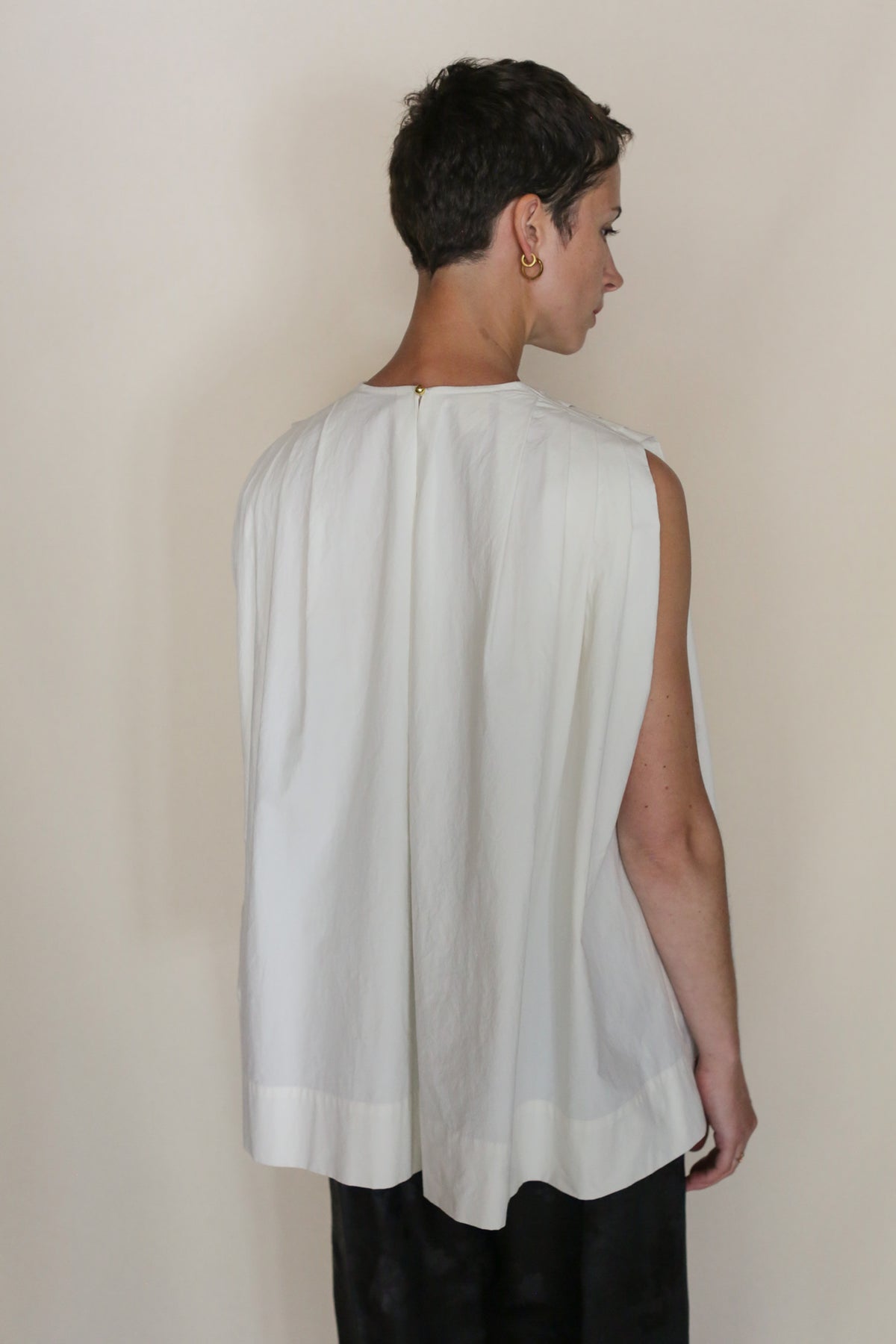 A person with short hair, wearing the Omizarashi Pleat Top and black pants, stands facing away from the camera against a neutral background.