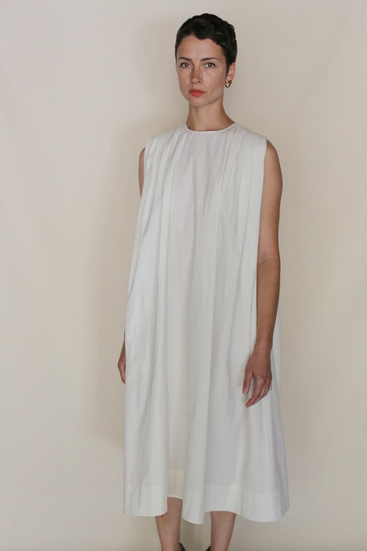 A person with short dark hair stands against a plain background, wearing the Omizarashi Pleat Dress—a loose, sleeveless white dress made from Ohmi Zarashi cotton. They look directly at the camera with a neutral expression.
