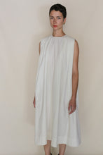 Load image into Gallery viewer, A person with short dark hair stands against a plain beige background, wearing the Omizarashi Undyed Shoulder Pleat Dress—a loose, sleeveless, knee-length white cotton dress—and gold earrings, looking at the camera with a neutral expression.