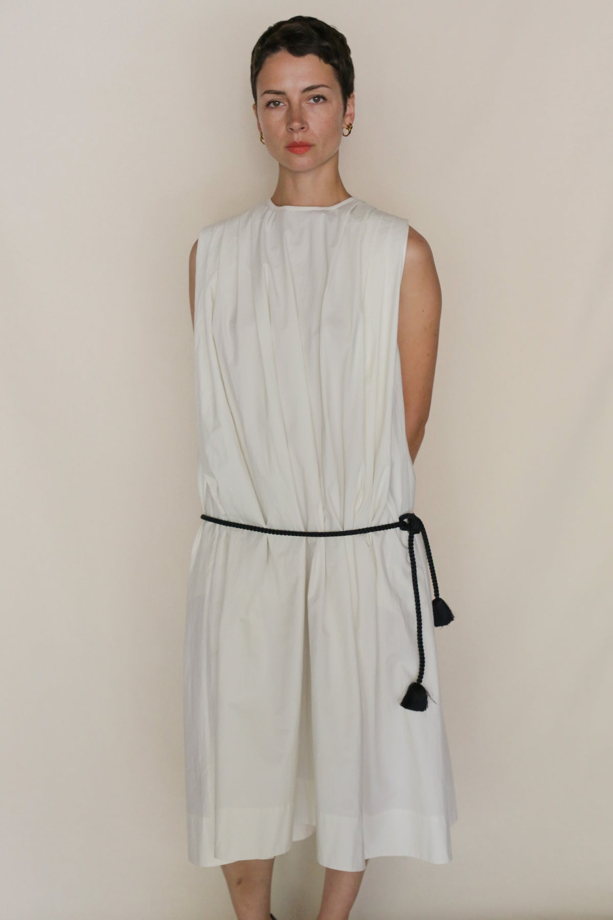 A person with short hair wears the Omizarashi Pleat Dress—sleeveless and loose-fitting with shoulder pleats—made from Ohmi Zarashi cotton, cinched with a black rope belt, and handmade in San Francisco against a beige background.