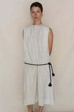 Load image into Gallery viewer, A person in the Shoulder Pleat Dress | Omizarashi Undyed—a sleeveless, loose white cotton dress with shoulder pleats and a black rope belt—stands against a plain beige background.
