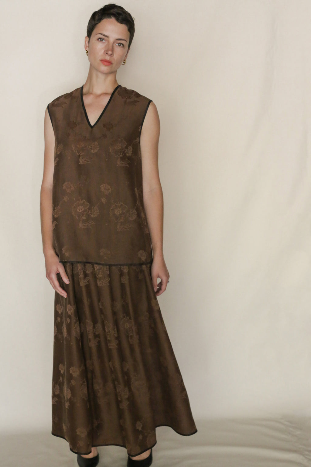 A woman with short dark hair wears the Lingering Fragrance Vest, a sleeveless loose brown silk jacquard dress with a subtle floral pattern, paired with black shoes and set against a plain light background.