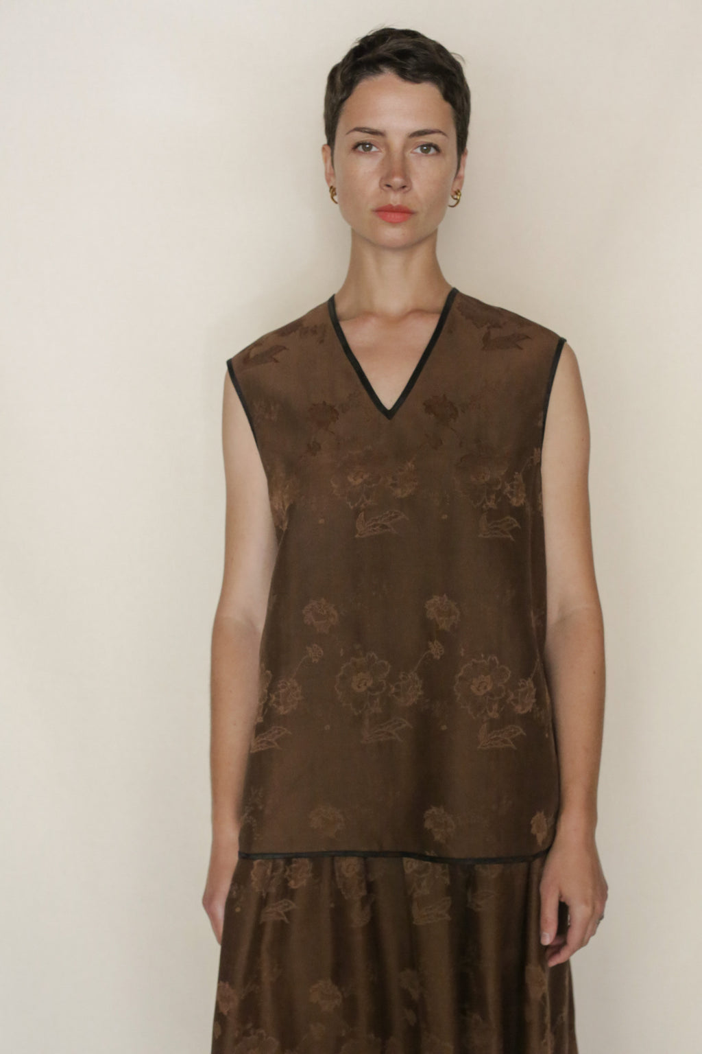 A person with short dark hair wears the Lingering Fragrance Vest, a sleeveless brown silk jacquard top with a subtle floral pattern and V-neck, standing against a plain light background.