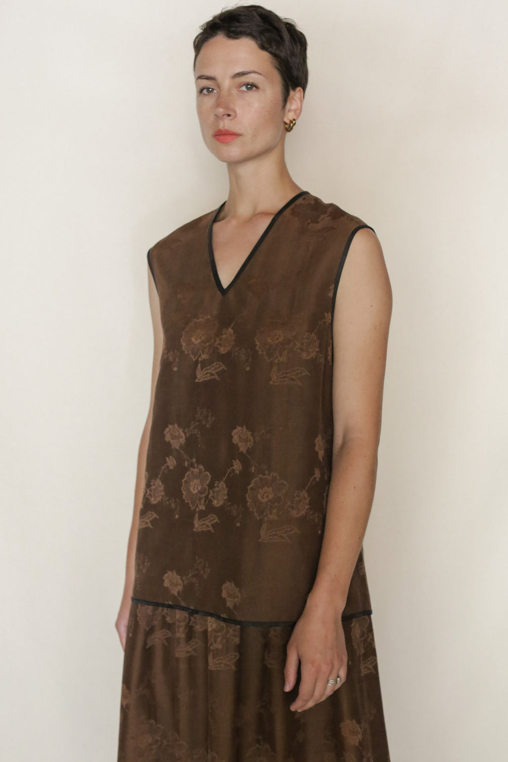 A person with short dark hair wears a sleeveless brown floral dress with a V-neck and black trim, paired with the Lingering Fragrance Vest in Xiang Yun Silk jacquard, gold hoop earrings, and stands against a plain light background.