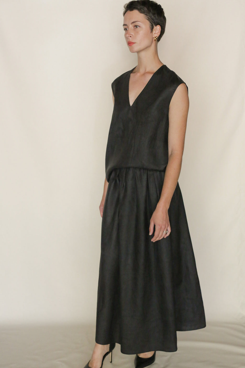 A woman with short hair wears a sleeveless black dress featuring the Faux Bois A Line Skirt and a V-neck, its long, flowing skirt draping elegantly. She stands against a plain light background, gazing slightly to the side.