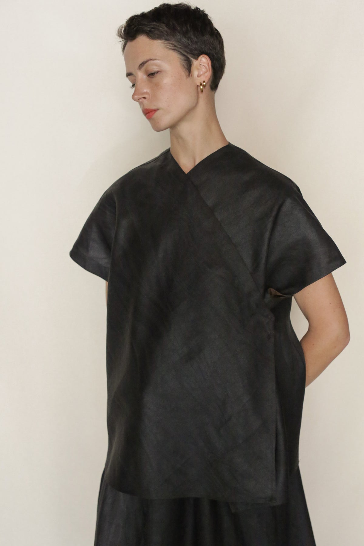 A person with short dark hair wears a loose black top styled like the Faux Bois Qipao Vest, featuring a wrap front. They stand against a plain light background, looking down with hands behind their back.
