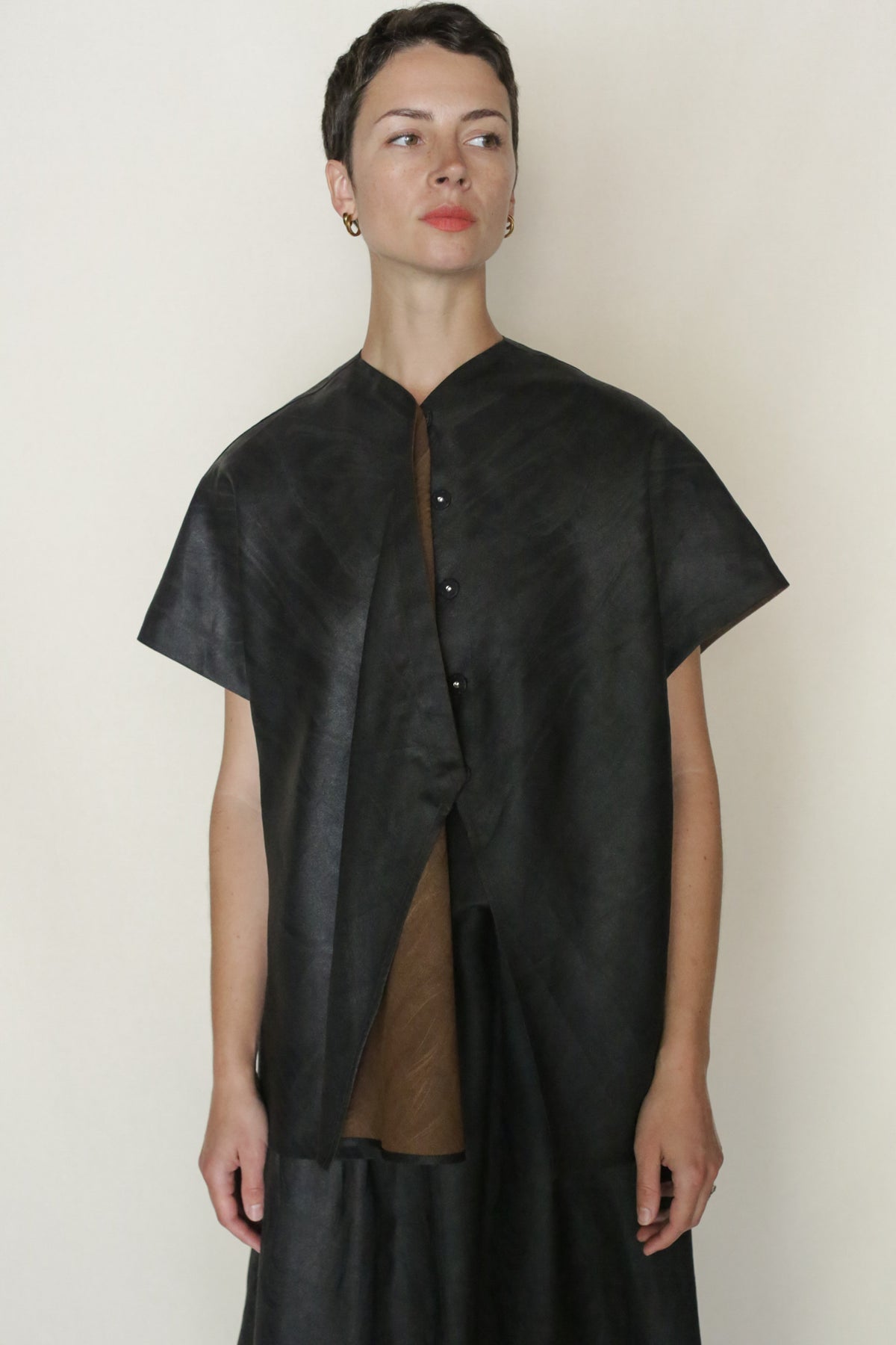 A person with short dark hair wears a black, short-sleeved, button-up top over a brown Faux Bois Qipao Vest, standing against an off-white background and looking slightly to the side.