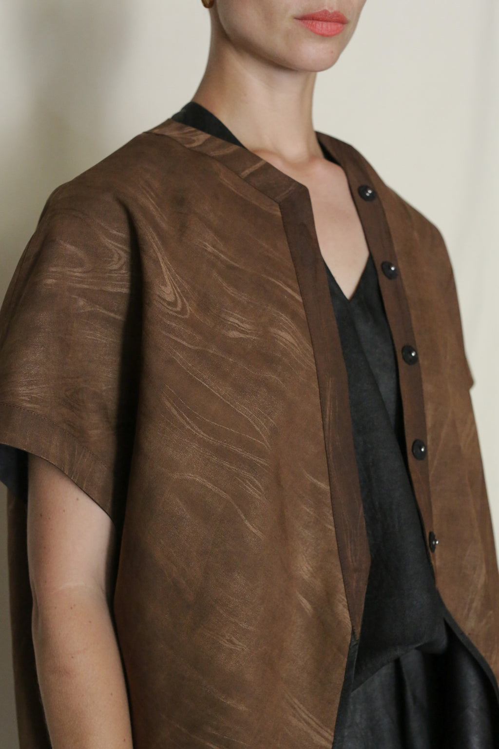 A person with short hair wears a loose, brown Faux Bois Qipao Vest with black buttons and a subtle wave pattern over a dark outfit, standing against a plain light background. Only the upper body is visible.