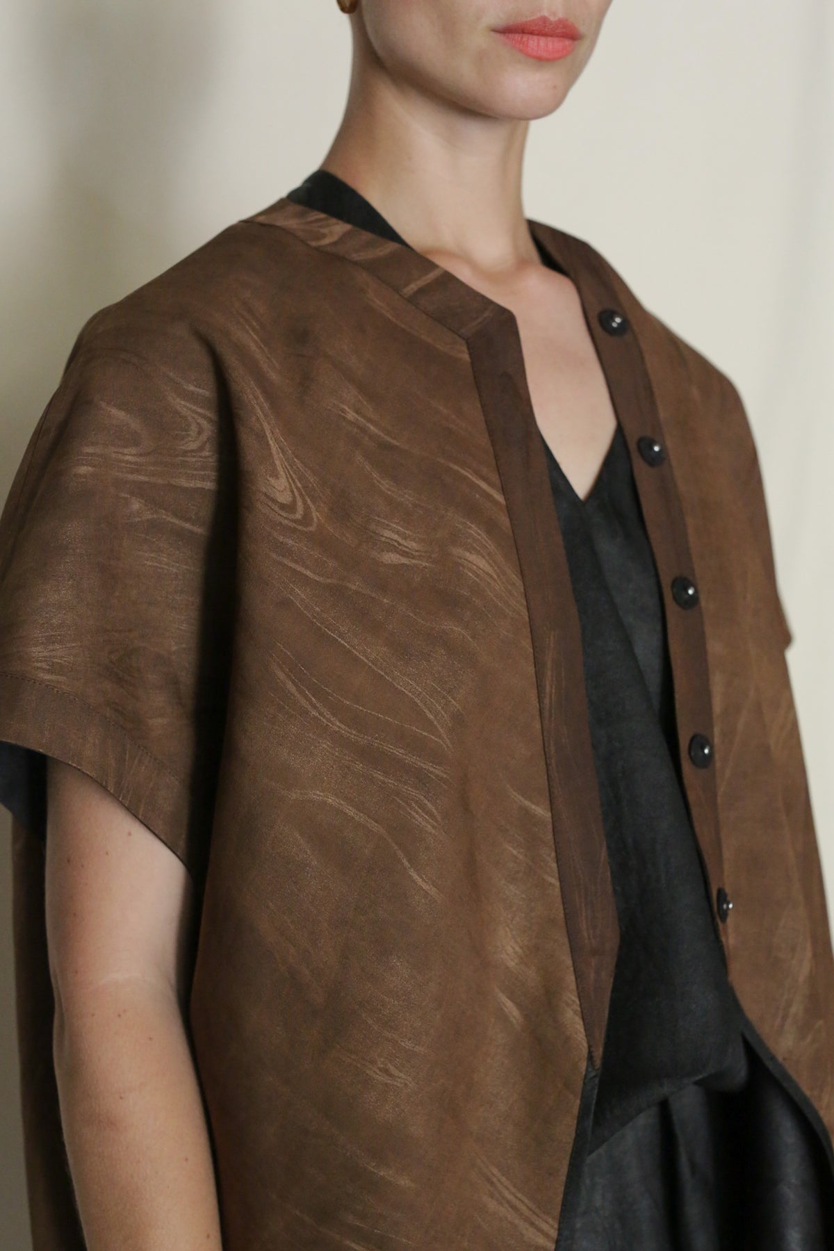 A person with short hair wears a loose, brown Faux Bois Qipao Vest with black buttons and a subtle wave pattern over a dark outfit, standing against a plain light background. Only the upper body is visible.